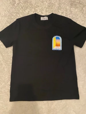 Svart Casablanca t-shirt med tryck - Svart t-shirt från Casablanca med färgglatt tryck på bröstet i blått, gult och orange. Klassisk rund hals och korta ärmar. Perfekt för dig som gillar stilrena plagg med en twist. Tillverkad i mjuk bomull.