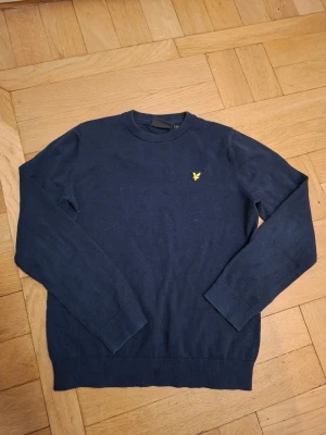 Mörkblå tröja från Lyle & Scott - Mörkblå långärmad tröja från Lyle & Scott med rund halsringning och gul broderad logga på bröstet. Tröjan har ribbade muddar vid ärmslut och nederkant. Perfekt för en stilren och enkel look.