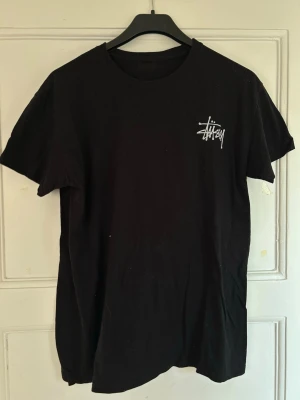 Svart Stussy t-shirt - Svart t-shirt från Stussy med vit logga på bröstet och stor tryckt Mickey Mouse-grafik på ryggen tillsammans med Stussy-tryck. Klassisk passform och rund hals. Perfekt för dig som gillar streetwear och unika prints.