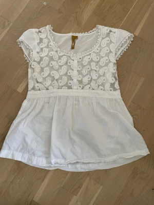 Vit spetsblus från Cream - Söt vit blus från Cream med spetsdetaljer och knappar framtill. Blusen har korta ärmar med spetskant och ett broderat mönster upptill. Perfekt för dig som gillar romantisk och feminin stil.