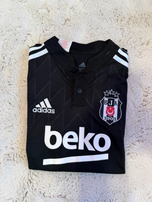 Svart Besiktas matchtröja Adidas - Snygg svart matchtröja från Besiktas med vita detaljer och klubbmärke på bröstet. Adidas-logga och sponsortryck på framsidan. Tröjan har korta ärmar, knappar vid halsen och är tillverkad i lätt funktionsmaterial.