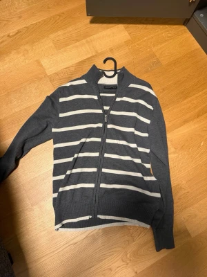 Gråvit randig ziptröja Selected Homme - Snygg ziptröja från Selected Homme i grått med vita horisontella ränder. Tröjan har hög krage, hel dragkedja och ribbade muddar. Perfekt för lager-på-lager och enkel att matcha med jeans eller chinos.