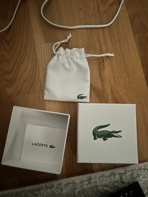 Vit presentask från Lacoste - Säljer en stilren vit presentask från Lacoste med matchande tygpåse och originalkort. Asken har Lacostes klassiska gröna krokodillogga på locket och på påsen. Perfekt för att ge bort accessoarer eller smycken.