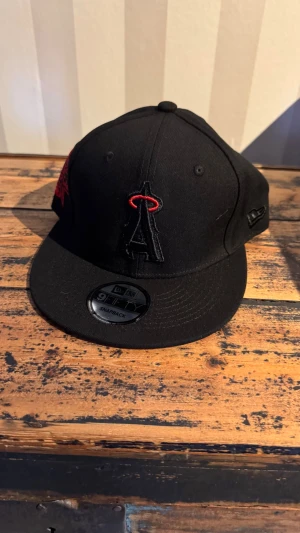 Svart New Era 9FIFTY snapback keps - Säljer en svart snapback keps från New Era, modell 9FIFTY. Kepsen har ett broderat A-logotyp framtill med röd detalj och en röd All-Star Game 2010 patch på sidan. Klassisk platt skärm och justerbar snapback baktill. Perfekt för dig som gillar sportig streetstyle.
