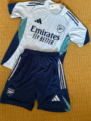  Arsenal fotboll set - Mörkblå sportiga short och t shirt från Adidas med Arsenal-logga på ena benet och klassiska vita Adidas-ränder på sidorna. Perfekta för träning eller fotboll, tillverkade i lätt och bekvämt polyestermaterial.