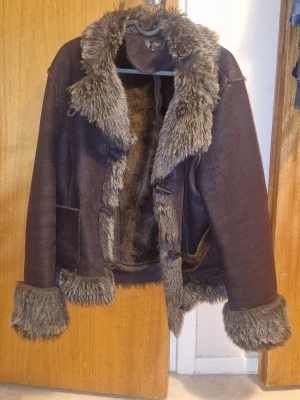 Jacka - Säljer en brun fin jacka i mjukt skönt material med fluffig fuskpäls i gråbrunt längs krage, ärmslut och nederkant. Jackan har två fickor-på ena har sömmen släppt lite men går att använda ändå och/eller enkel att fixa, jackan stängs med snygga knappar och öglor. Perfekt för dig som vill ha en trendig look.