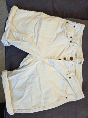 Vita shorts från Jack & Jones - Säljer ett par vita shorts från Jack & Jones i bomull. De har klassisk femficksdesign, knappar i gylfen och upprullade benslut. Perfekta för varma dagar och enkel att matcha med olika stilar.