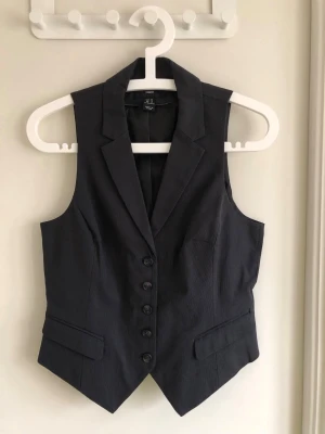 Svart kostymväst från H&M - Stilren svart kostymväst från H&M med klassisk krage och knäppning framtill. Västen har två fickor med lock och ett justerbart spänne i ryggen för perfekt passform. Perfekt att styla över en skjorta eller t-shirt för en trendig look.
