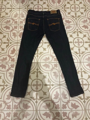 Nudie Jeans - Säljer ett par mörkblå jeans från Nudie Jeans med klassisk gul kontrastsöm. De har knappt inga tecken på användning!!  Längd: 97cm.  Bredd: 40cm