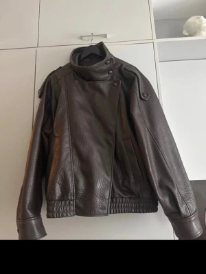 Mörkbrun skinnjacka med hög krage - Säljer en mörkbrun skinnjacka med hög krage och snygga knappdetaljer vid halsen från zara. Jackan har breda muddar i midjan och vid ärmsluten samt en cool asymmetrisk design framtill. Perfekt för dig som vill ha en unik och trendig look.