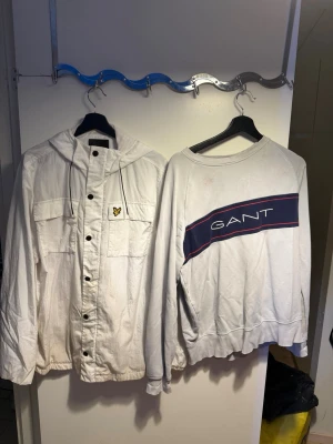 Vit vindjacka med huva från Lyle & Scott Gant Swertshirt Säljs som Paketpris! - Hej säljer en Vit vindjacka från Lyle & Scott med huva, snörning och tryckknappar framtill. Jackan har två stora bröstfickor och en gul logotyp på vänster sida. Perfekt för dig som gillar stilren och sportig look.