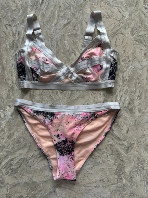Rosa och svart bikini set med silvriga band - Snyggt tvådelad bikini med bh och trosa. Färgen är ljusrosa med svarta och vita splash-detaljer och silvriga band. Bh:n har dubbla axelband och dekorativa band över kuporna. Perfekt för dig som gillar unika detaljer. OBS: FRAKTAR INOM 24H!🩷