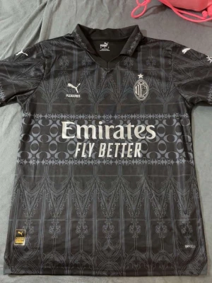Svart AC Milan matchtröja Puma - Säljer en svart AC Milan matchtröja från Puma med detaljerat mönster och klubbmärke på bröstet. Tröjan har korta ärmar, krage och tryckta loggor från Puma, Pleasures och Emirates Fly Better. Materialet är lätt och andas, perfekt för fotboll eller träning.