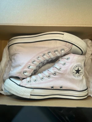 Ljusrosa Converse All Star High - Säljer ett par klassiska Converse All Star high sneakers i ljusrosa canvas med vita gummisulor och svarta detaljer. Skorna har den ikoniska loggan på sidan, vita skosnören och metallfärgade öljetter. Perfekta för en avslappnad och trendig stil.