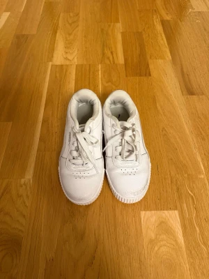 Vita Puma sneakers i läder - Snygga vita sneakers från Puma i klassisk låg modell. Skorna är tillverkade i läder med matchande vita skosnören och diskret Puma-logga på sidan. Perfekta för en clean och stilren look till vardagsoutfiten.