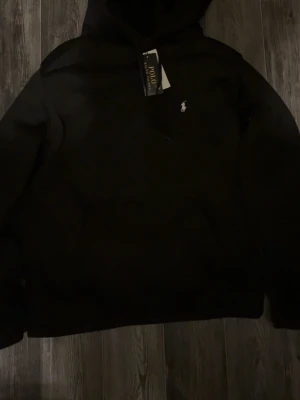 Svart hoodie från Polo Ralph Lauren - Snygg svart hoodie från Polo Ralph Lauren med klassisk huva och den ikoniska lilla loggan broderad på bröstet. Tillverkad i mjukt material som känns skönt mot huden. Perfekt för dig som vill ha en stilren och enkel look.