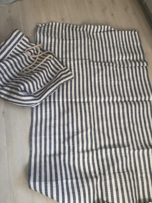 Randig shoppingväska med rep-handtag - Säljer en stor shoppingväska i tyg med marinblå och vita ränder. Väskan har rejäla rep-handtag och en mörkblå insida. Perfekt för stranden eller shoppingrundan när du vill ha plats för mycket. Klassisk och stilren design.Handtsgen på väskan är lagade men håller ett bra tag till, filten oanvänd 