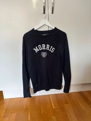 Marinblå tröja från Morris - Snygg marinblå tröja från Morris med vit broderad logga och emblem på bröstet. Tröjan har rund halsringning, långa ärmar och ribbade muddar vid ärmslut och nederkant. Perfekt för en stilren och avslappnad look. I storlek S.