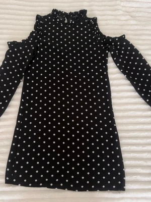 Svart prickig klänning med volang - Snygg svart kortklänning med vita prickar och volangdetaljer vid axlar och hals. Klänningen har lång ärm och en hög krage med rynk. Perfekt för dig som gillar en lekfull men stilren look.