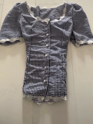 Blårutig blus med spetsdetaljer vintage - Supersöt blus i blåvitt rutigt mönster med korta ärmar och vita spetsdetaljer vid halsringning, ärmslut och nederkant. Knäpps framtill med pärlknappar och har en feminin, figurnära passform. Perfekt för en trendig och söt look.