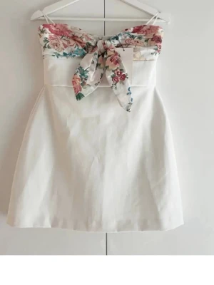 Vit axelbandslös klänning med rosett - Söt axelbandslös kortklänning i vitt med blommigt mönster och stor rosett upptill. Klänningen har en feminin siluett och är perfekt för sommarens alla tillfällen. 