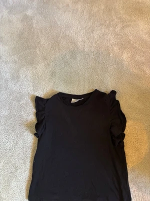 Svart topp med volangärmar i strl 146/152 från Gina Tricot  - En svart topp med rund hals och volangdetaljer vid ärmarna. Topprn har en enkel, stilren design som passar till mycket. Tillverkad i mjukt material för en bekväm känsla.