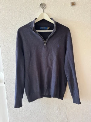 Mörkblå stickad tröja Polo Ralph Lauren - Mörkblå stickad tröja från Polo Ralph Lauren med halv dragkedja och diskret broderad logga på bröstet. Tröjan har lång ärm, ribbade muddar och hög krage. Perfekt för lager på lager och klassisk stil.