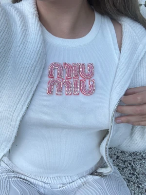 Miu Miu linne - Snygg vit ribbad linnetopp med breda axelband och ett coolt pärlbroderat motiv i rosa och vitt framtill. Toppen har en croppad passform och är perfekt för varma dagar. Materialet känns mjukt och stretchigt.