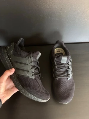 Svarta Adidas Ultraboost sneakers - Säljer ett par svarta Adidas Ultraboost sneakers med stickad ovandel och klassiska tre ränder på sidan. Skorna har svart sula och snörning, samt diskret Adidas-logga på hälen. Perfekta för dig som gillar stilrena och bekväma sneakers. Priset är diskuterbart vid snabbt köp. Skickar fort! Tveka inte att skicka och fråga om du har några frågor!