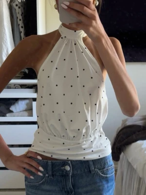 Vit prickig halterneck topp - Säljer en vit halterneck topp med svarta prickar och resår i midjan. Toppen är ärmlös och har en hög hals som knyts i nacken. Perfekt till jeans eller kjol för en trendig look. Stl xs, helt ny! Från SHEIN 