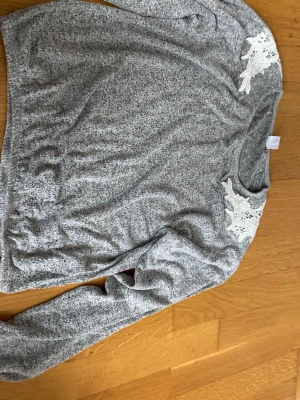 Gråmelerad stickad tröja med spets - Mysig gråmelerad stickad tröja med långa ärmar och rund hals. Tröjan har vita spetsdetaljer på axlarna som ger en extra touch. Perfekt för dig som gillar en enkel men ändå unik stil. Materialet känns mjukt och bekvämt.