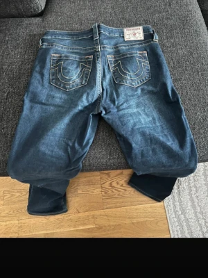 Blå True Religion jeans med kontrastsömmar - Säljer ett par klassiska blå jeans från True Religion med ikoniska kontrastsömmar på bakfickorna och patch med logga i midjan. Jeansen har rak passform, fem fickor och är tillverkade i denim med snygg tvätt. Perfekta för en avslappnad och trendig stil.