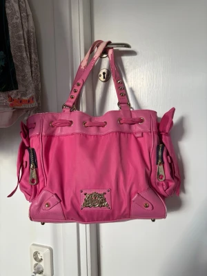 Juicy Couture rosa handväska - Säljer en stor rosa handväska från Juicy Couture med två axelband, dekorativa nitar och dragkedjor på sidorna. Väskan har en metallbricka med logga framtill och detaljer i guld. Perfekt för dig som gillar färgstarka accessoarer.