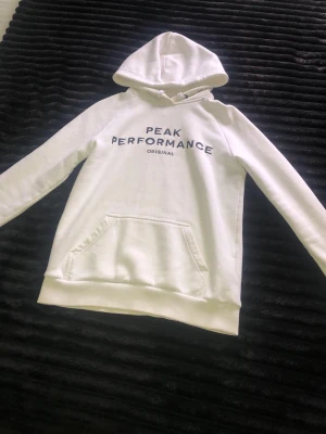Vit hoodie från Peak Performance - Snygg vit hoodie från Peak Performance med svart text på bröstet. Klassisk huva med dragsko och stor känguruficka framtill. Tillverkad i mjukt material som känns skönt mot huden. Perfekt för en avslappnad och sportig stil.