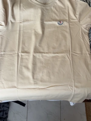 Beige t-shirt från Moncler - Snygg beige t-shirt från Moncler med klassisk rund halsringning och liten Moncler-logga på bröstet. Tillverkad i mjuk bomull som känns skön mot huden. Perfekt basplagg för en clean och stilren look.