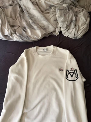 Vit sweatshirt från Moncler - Säljer en stilren vit sweatshirt från Moncler med rund halsringning. Tröjan har två broderade Moncler-loggor på bröstet och ärmen samt en stor svart M-detalj på ärmen. Perfekt för dig som gillar exklusiva streetwear-plagg.