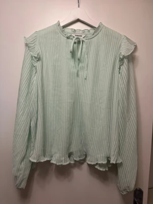 Ljusgrön blus med volanger - Söt ljusgrön blus från Kappahl med plisserat tyg, volangdetaljer vid axlar och nederkant samt knytband i halsen. Blusen har långa ärmar och en luftig, flowy passform som ger en romantisk vibe. Perfekt för dig som gillar en feminin och trendig stil.