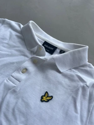 Vit pikétröja från Lyle & Scott - OBS❗️ALLT KOMMER ATT STRYKAS INNAN FRAKT! Klassisk vit pikétröja från Lyle & Scott med korta ärmar, krage och knappar framtill. Tröjan har det ikoniska gula logomärket broderat på bröstet. Perfekt till en clean och stilren look. Passar dem runt 165-175. Inga synliga defekter