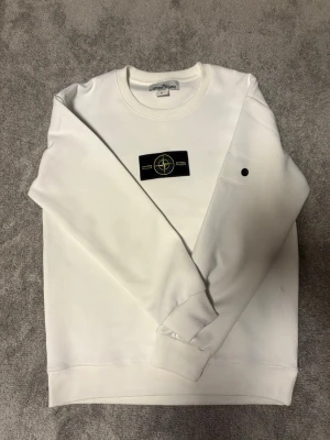 Vit sweatshirt från Stone Island - Vit sweatshirt från Stone Island med klassisk rund hals och ribbade muddar. Tröjan har en svart patch med logga på bröstet och är tillverkad i mjukt bomullsmaterial. Perfekt för en clean och stilren look.