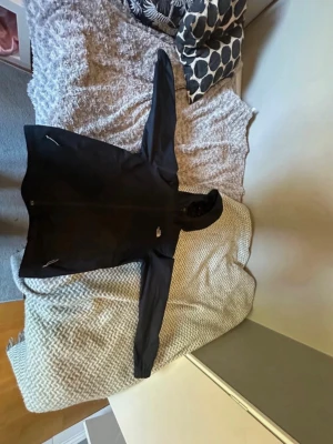 Svart vindjacka från The North Face - Svart vindjacka med huva från The North Face. Jackan har dragkedja framtill, två sidofickor med dragkedja och meshfoder på insidan. Perfekt för blåsiga dagar och enkel att bära med sig.