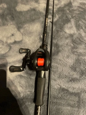 Svart baitcasting fiskespö med rulle - Säljer ett svart baitcasting fiskespö med matchande rulle och röd lina. Handtaget är i mjukt gummimaterial för bra grepp och spöet har en kolfiberlook. Perfekt för dig som gillar sportfiske och vill ha ett stabilt och snyggt spö.