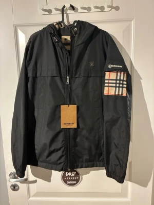 Svart vindjacka från Burberry - Snygg svart vindjacka från Burberry i storlek small med dragkedja framtill, huva och klassiskt rutigt Burberry-mönster på insidan och på ena ärmen. Jackan har en modern passform och diskret Burberry-logga på bröstet. Perfekt för dig som vill ha en stilren och ikonisk look.