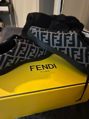Fendi sneakers med FF-mönster - Snygga sneakers från Fendi med ikoniskt FF-mönster i svart och grått. Skorna har en låg profil, svarta sulor och svarta snören. Materialet är textil med detaljer i läder. Perfekt för dig som vill sticka ut med exklusiva designer.