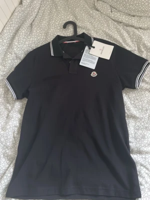 Svart pikétröja från Moncler - Svart pikétröja från Moncler med klassisk krage och vita ränder på krage och ärmslut. Tröjan har Moncler-logga broderad på bröstet och knappar vid halsen. Perfekt för en stilren och sportig look.