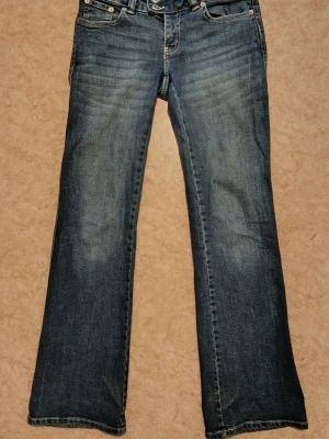 Blå bootcut jeans med broderade fickor - Säljer ett par klassiska blå bootcut jeans med snygg slitning framtill och broderade detaljer på bakfickorna. Jeansen har normal midja och fem fickor. Perfekta för dig som gillar en avslappnad men ändå trendig stil.