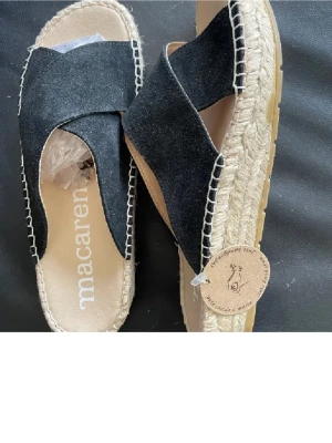 Svarta sandaler från Macarena - Snygga svarta sandaler från Macarena med breda korslagda remmar i mocka och naturfärgad flätad sula. Yttersulan är i gummi för bra grepp. Perfekta för en avslappnad och trendig look under sommaren. Nya oanvända 