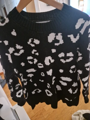 Svartvit leopardmönstrad stickad tröja - Stickad tröja i svart med vitt leopardmönster. Tröjan har rund halsringning och ribbade muddar vid ärmslut och nederkant. Perfekt för dig som gillar djurmönster och vill ha en trendig look.