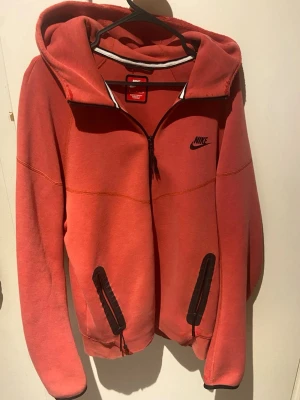 Nike tech fleece röd - Röd hoodie från Nike med dragkedja framtill och två svarta fickor med dragkedja. Klassisk Nike-logga på bröstet och stor huva. Tillverkad i mjukt bomullsmaterial som är skönt mot huden. Perfekt för en avslappnad streetwear-look.