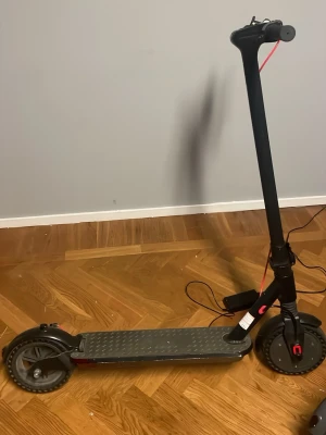 Svart elscooter med display - Elektrisk scooter i svart med digital display på styret, grått halkskydd på fotplattan och röda detaljer. Scootern har stora hjul, handbroms och laddare medföljer. Perfekt för smidig transport i stan. 