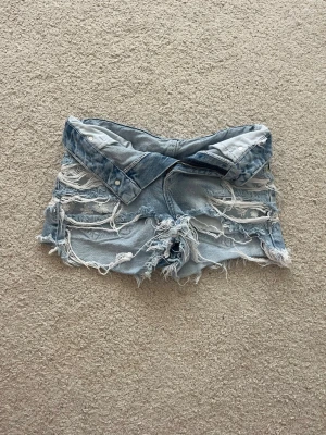 Ljusa denimshorts från Bershka - Säljer ett par ljusblå denimshorts från Bershka med låg midja och slitna detaljer. Shortsen har råa kanter och flera revor framtill för en edgy look. Perfekta till sommaren och ger en avslappnad vibe.